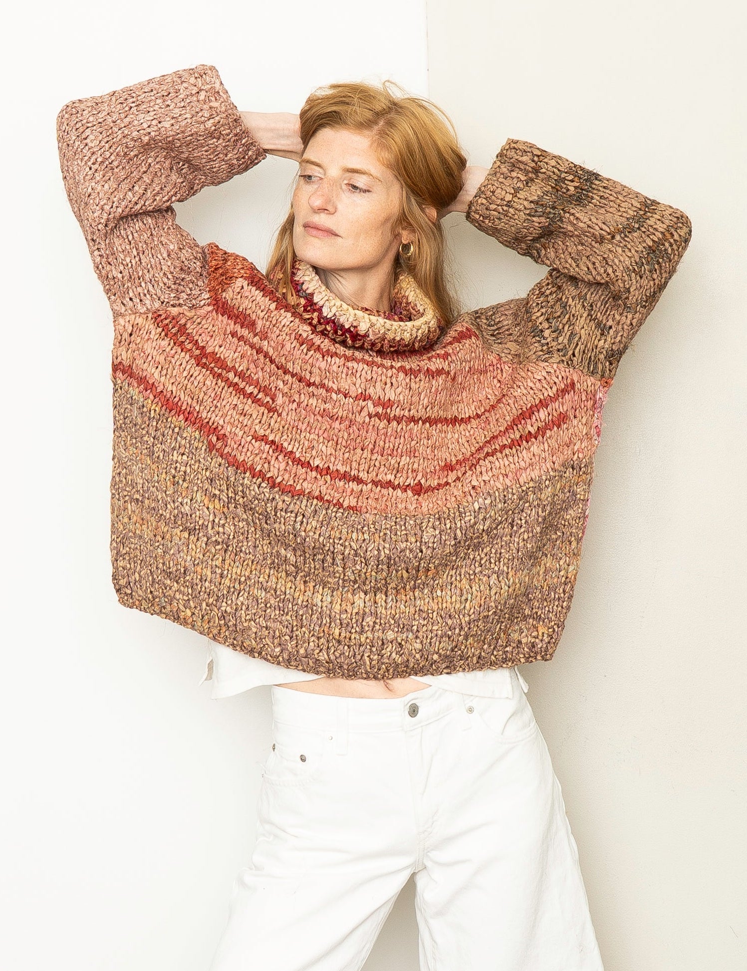 A woman wearing a chunky knit turtleneck sweater with horizontal stripes in brown, rust red, and beige, paired with white shorts, posing against a plain off-white wall.