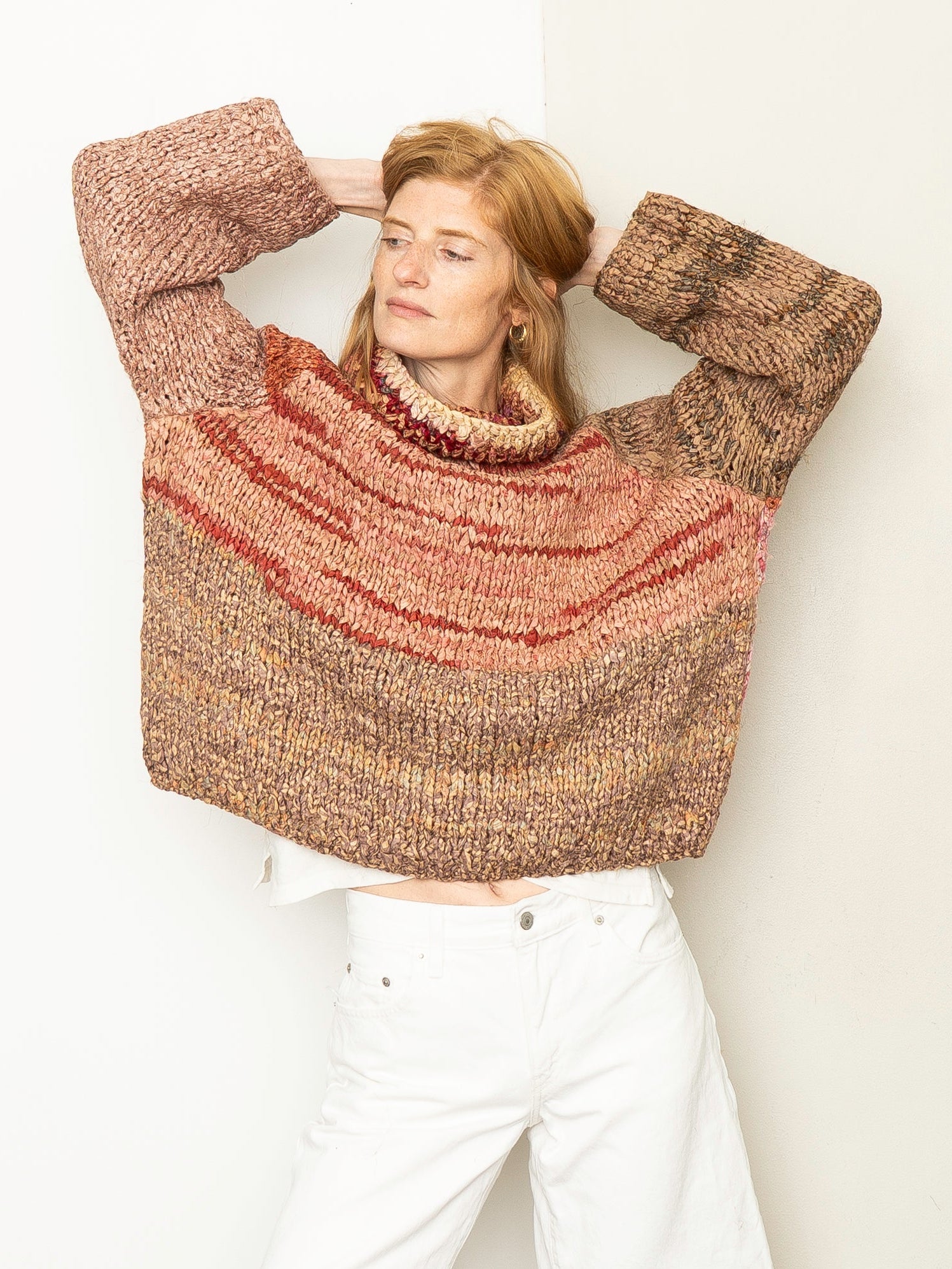 A woman wearing a chunky knit turtleneck sweater with horizontal stripes in brown, rust red, and beige, paired with white shorts, posing against a plain off-white wall.